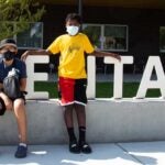 Two students stand together outside wearing masks in front of a school