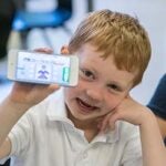 A young student sits in a classroom holding a mobile phone and smiling for a photo.