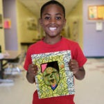 A young student holds up their art work and smiles for a photo in a school hallway