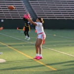 Two women's flag football teams play a match on an athletic field. A ref moves in to watch the play