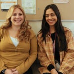 Two women sit together on a couch and smile for a photo