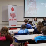 A group of students sit in a conference room with a student panel at the front of the room. An adults provides group facilitation.