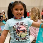 A elementary student smiles as she holds a bag with books in a school auditorium