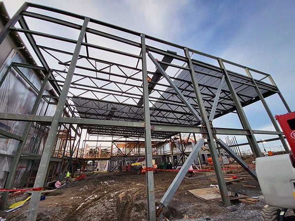 a structural steel frame for a building with an existing building tarped off next to it