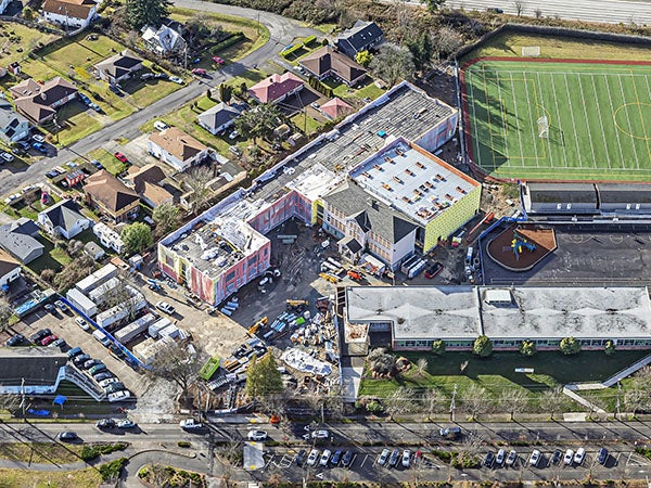 aerial of large building under construction, another large building at the lower right, and a green turf athletic field
