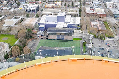 a curved orange surface in the foreground looks down to an athletic field and grandstands below with other buildings on the outside