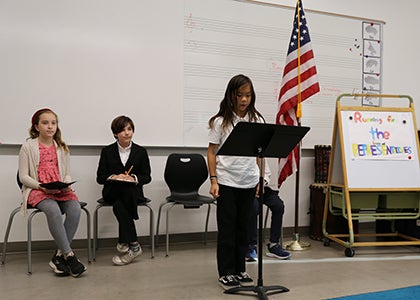 A student with two students behind them stand in front of a classroom at a music stand