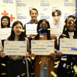 A group of eight students hold large checks while confetti falls on them