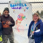 Four adults stand by a banner created by students on a school campus.