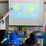 Two student teachers stand before elementary age students at a map of the world