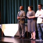 A small group of people stand together on a stage with one student at a microphone and Dr. Jones and School Board President Rankin watching.