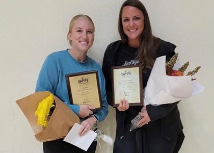Two women stand together with flowers and awards