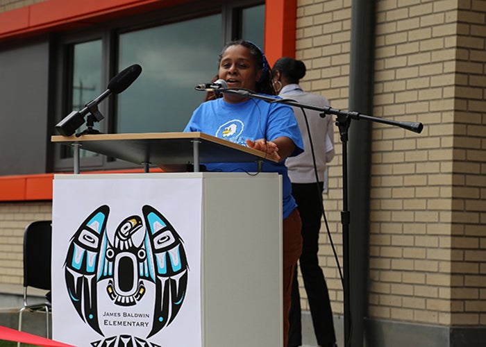 a woman stands at a podium with microphones attached. a light colored brick wall and a window with an orange frame are behind her