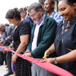 a group of people are looking down at a red ribbon. one person has giant scissors. the others have normal size scissors. all have the ribbon between the scissor blads