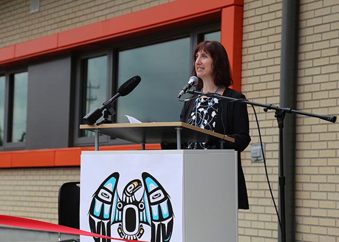 a woman stands at a podium with a microphone attached. a light colored brick wall and a window with an orange frame are behind her