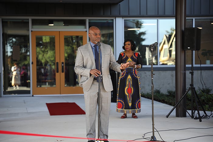 a man wearing a suite stands at a microphone. a woman in a dress stands behind him. both are in front of large glass doors.