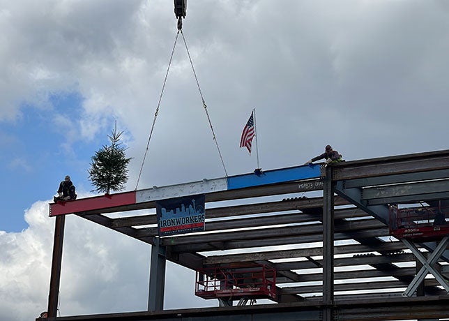 2 people on top of a steel structure on each side of a red, white and blue beam that has an american flag and an evergreen tree on it