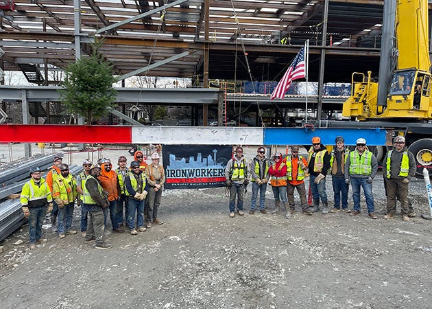a group of people in construction safety vests and hats stand in front of a steel beam with a steel framed structure behind