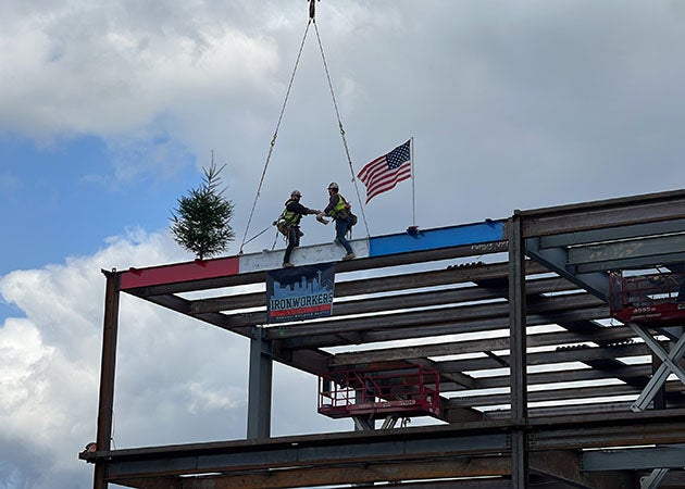 2 people stand on a red, white, and blue beam on one corner of a steel framed structure. they are shaking hands. an evergreen tree is on one end and a United States flag is on the other.