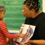 A teacher and preschool student read a book together.