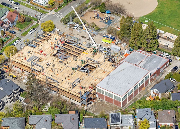 Aerial view of a school construction site with a wooden and steel-framed building next to a red-brick gym, surrounded by homes, a playground, and a sports field.