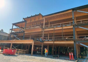 Ground-level view of a multi-story school building under construction with exposed wood, steel framing, and workers on-site.