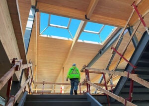 View up a stairwell where a worker walks toward a skylight-filled wooden ceiling under construction.
