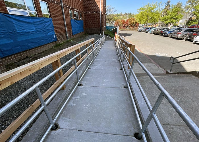 A newly constructed concrete accessibility ramp with metal railings leads to the side entrance of a red-brick school, with a parking lot visible in the background.