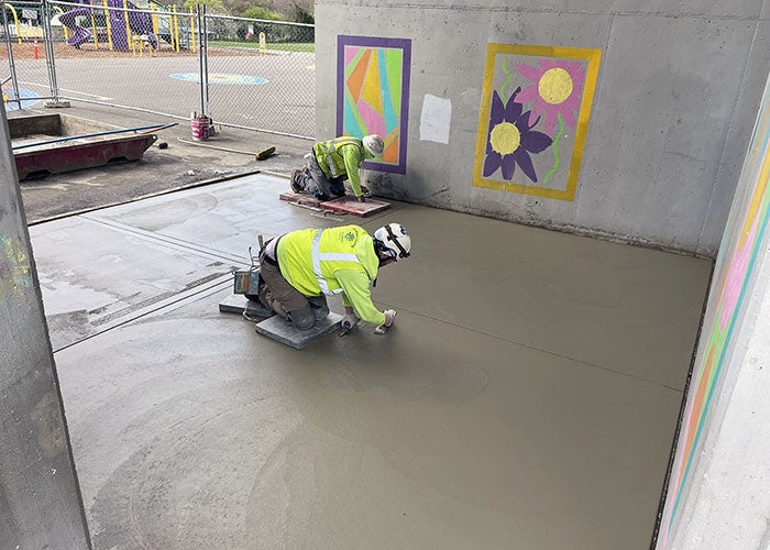 Two construction workers smooth fresh concrete beneath a structure decorated with colorful murals, near a fenced playground area.