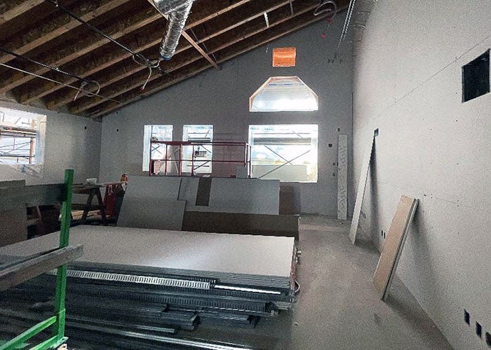A room with a pitched ceiling and exposed trusses shows drywall installation in progress, with light streaming through tall windows.