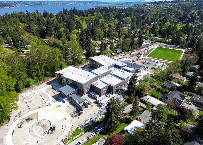 Aerial shot of a large, modern school campus surrounded by trees and residential neighborhoods, with construction equipment visible and a finished turf field in the distance.