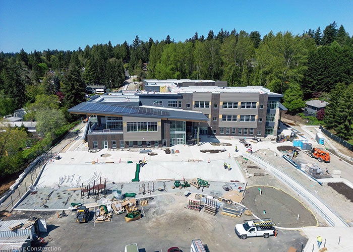 Ground-level view of the front of the school building, showing large windows, solar panels on the roof, and active construction around the entry plaza.