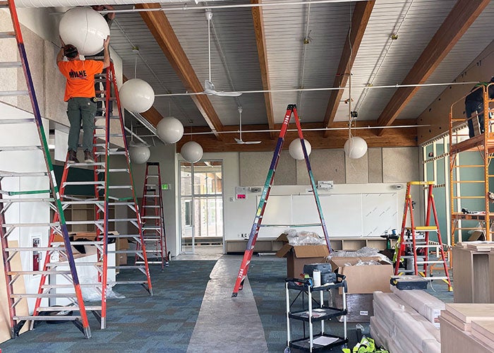 Library scene with ladders, workers installing large round light fixtures, and ceiling beams exposed above.