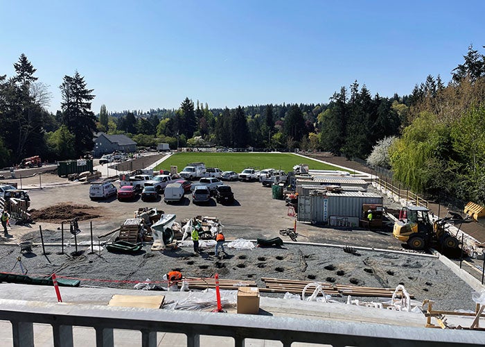 Construction crews working near the edge of a turf field, with parked vehicles and containers on-site and a forested backdrop.