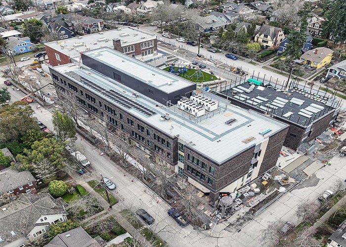 Overhead view showing a nearly complete modern school addition next to a historic red-brick school building, with visible rooftop units, solar panels, and surrounding neighborhood streets.