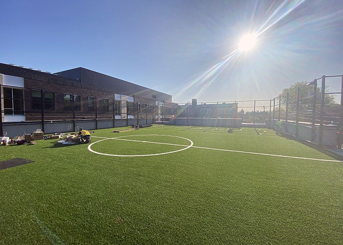 A rooftop turf field with sports markings, surrounded by safety fencing, under a clear blue sky with bright sunlight.