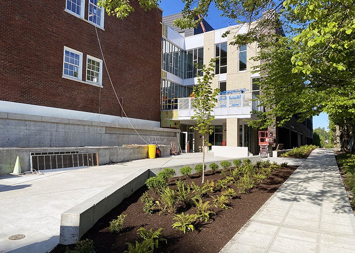 A planted entry area with new shrubs, young trees, and a concrete walkway connecting the original red-brick school with the modern expansion.
