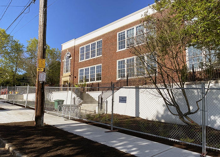 View of the original red-brick school facade with a freshly poured sidewalk, new fencing, and a terraced landscape leading to the entrance.