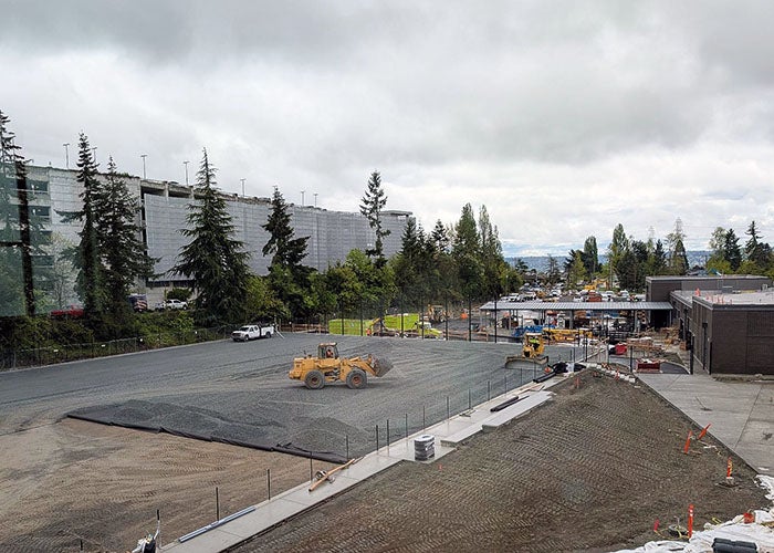 View of an active construction site where heavy equipment is grading a large gravel area, with adjacent buildings, fencing, and a parking structure in the background.