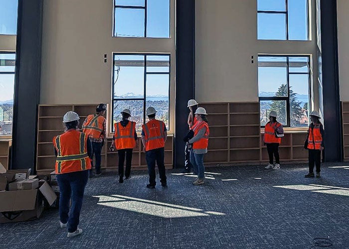 A group of construction workers and visitors in hard hats and safety vests stand in a bright room with tall windows and built-in shelving.