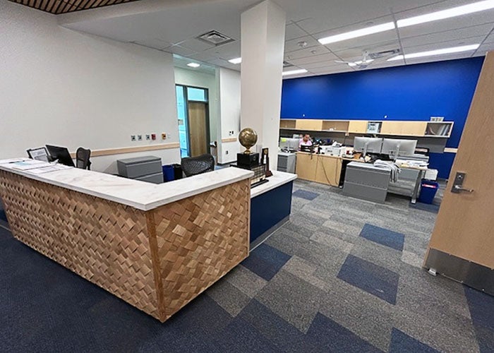 nterior view of a school office featuring a wood-accented reception desk and blue wall backdrop, with office cubicles in the background.
