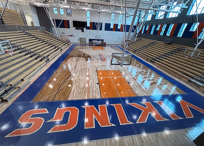 Full gymnasium with polished wood floors, “VIKINGS” lettering, bleachers on both sides, and high ceilings with natural light.