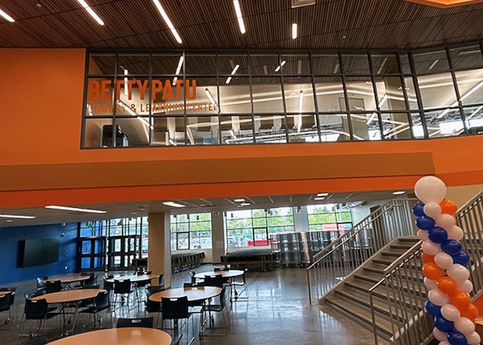 View of the school's commons with round tables, balloons, and large windows. “Betty Patu Library & Learning Center” is displayed above.
