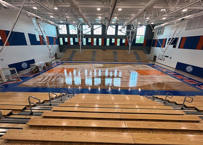 View from the top of the bleachers looking across the full gym floor with basketball court lines and team branding visible.