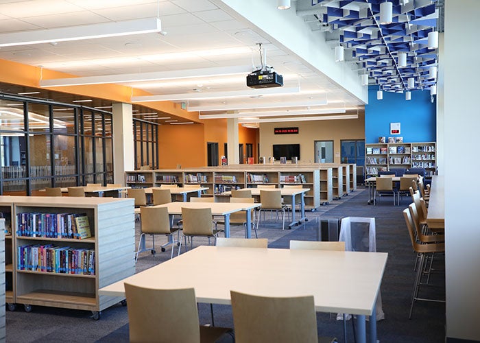 Additional view of the library with study tables, bookshelves, orange and blue accent walls, and modern ceiling design.