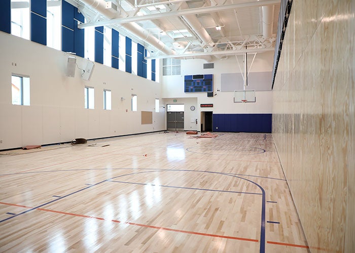 Bright, smaller gym space with white walls, light wood flooring, a scoreboard, and blue acoustic paneling.