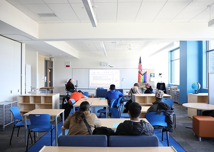 Students and a teacher gather in a modern, flexible classroom with blue chairs, modular tables, and a digital projector screen at the front.
