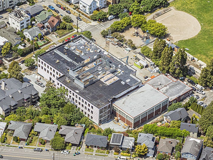 Aerial view of a large school or institutional building under renovation, surrounded by residential homes. The flat roof is partially completed, with construction materials and equipment visible on top. Adjacent to the building is a playground and green field, and construction activity is ongoing in the surrounding lot