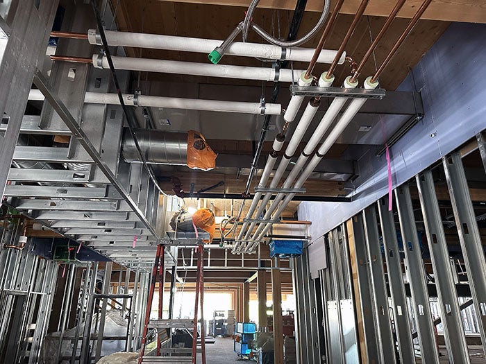Interior shot of a building under construction, showing exposed ceiling framing with newly installed ducts and piping. Electrical conduits and plumbing pipes are neatly aligned overhead, while the floor below is filled with framing studs and construction equipment.