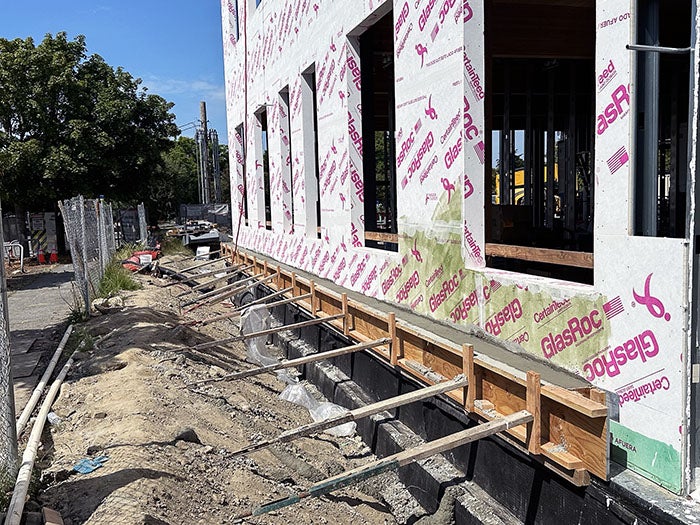 Exterior view of a building wrapped in GlasRoc sheathing, with foundation work in progress. Wooden forms are set up for concrete pouring along the base of the wall, and soil and construction debris line the ground alongside the building.